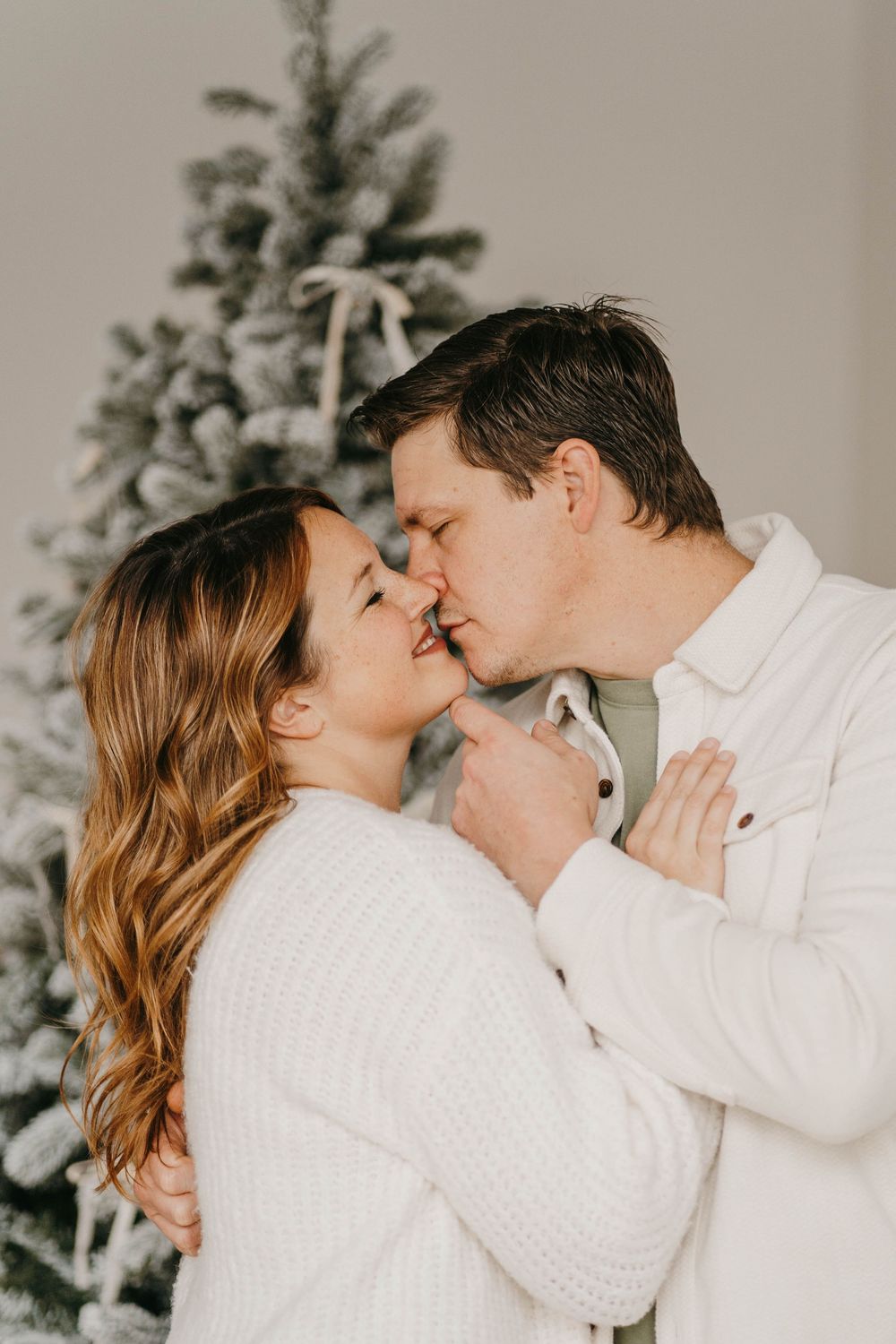 A couple in white sweaters share a romantic kiss in front of a snow-flocked Christmas tree.