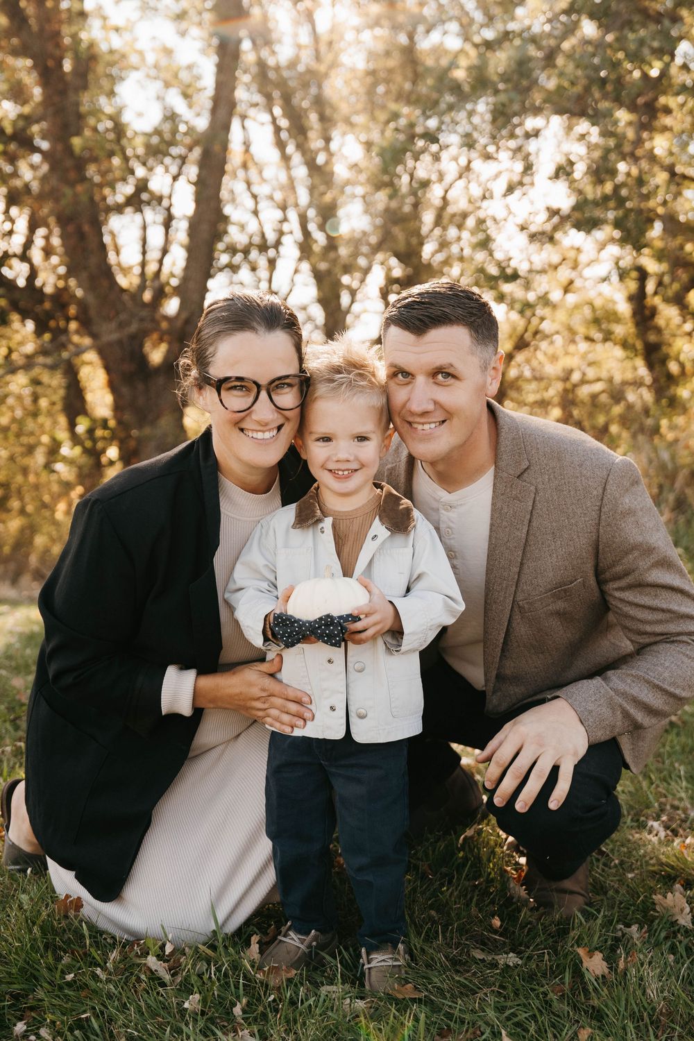 Family portrait of parents and young child in bow tie posing outdoors in golden sunlight among trees.