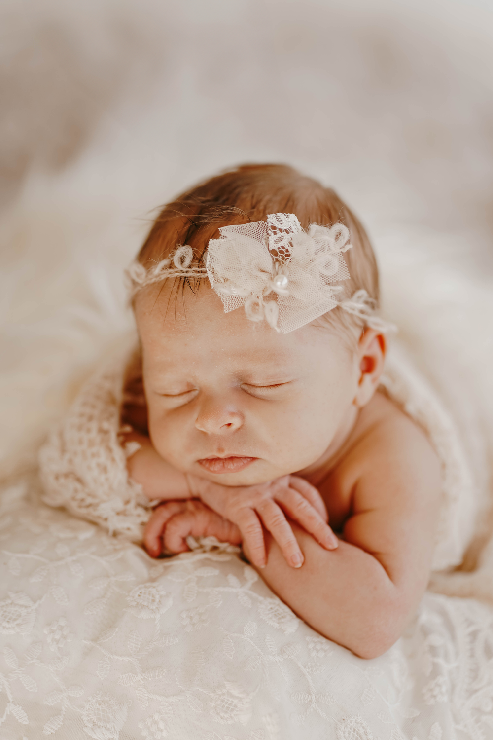 Sleeping newborn baby girl wearing a delicate white lace headband with bow, resting on soft white lace fabric.