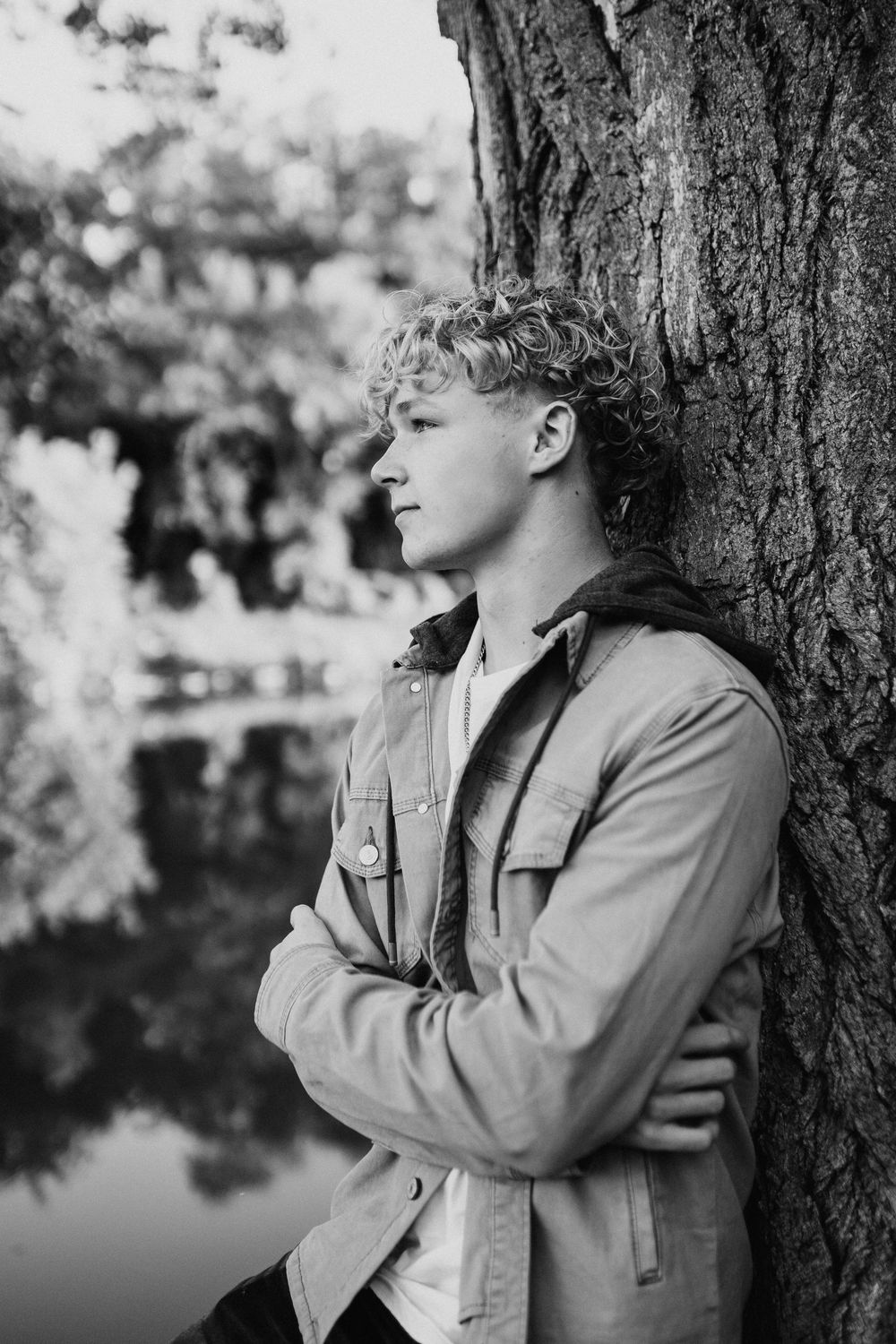 Black and white portrait of a young curly-haired man leaning against a tree with arms crossed outdoors.