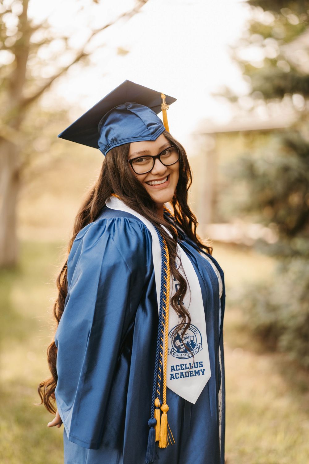 Smiling female graduate in blue cap and gown with Acellus Academy stole poses outdoors in warm sunlight.