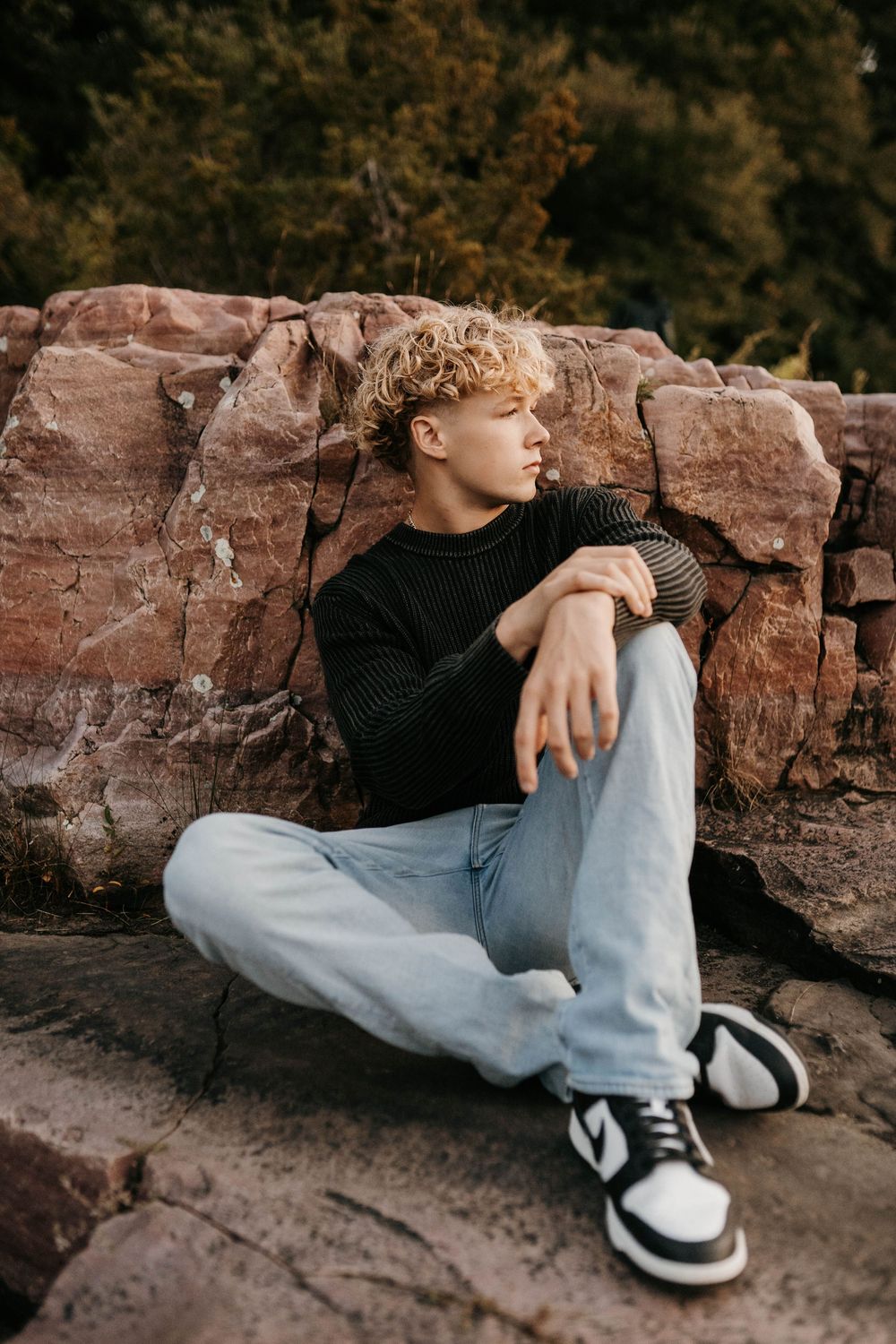 Young man with curly blonde hair sits against red rocks wearing black sweater, light jeans, and Nike sneakers.