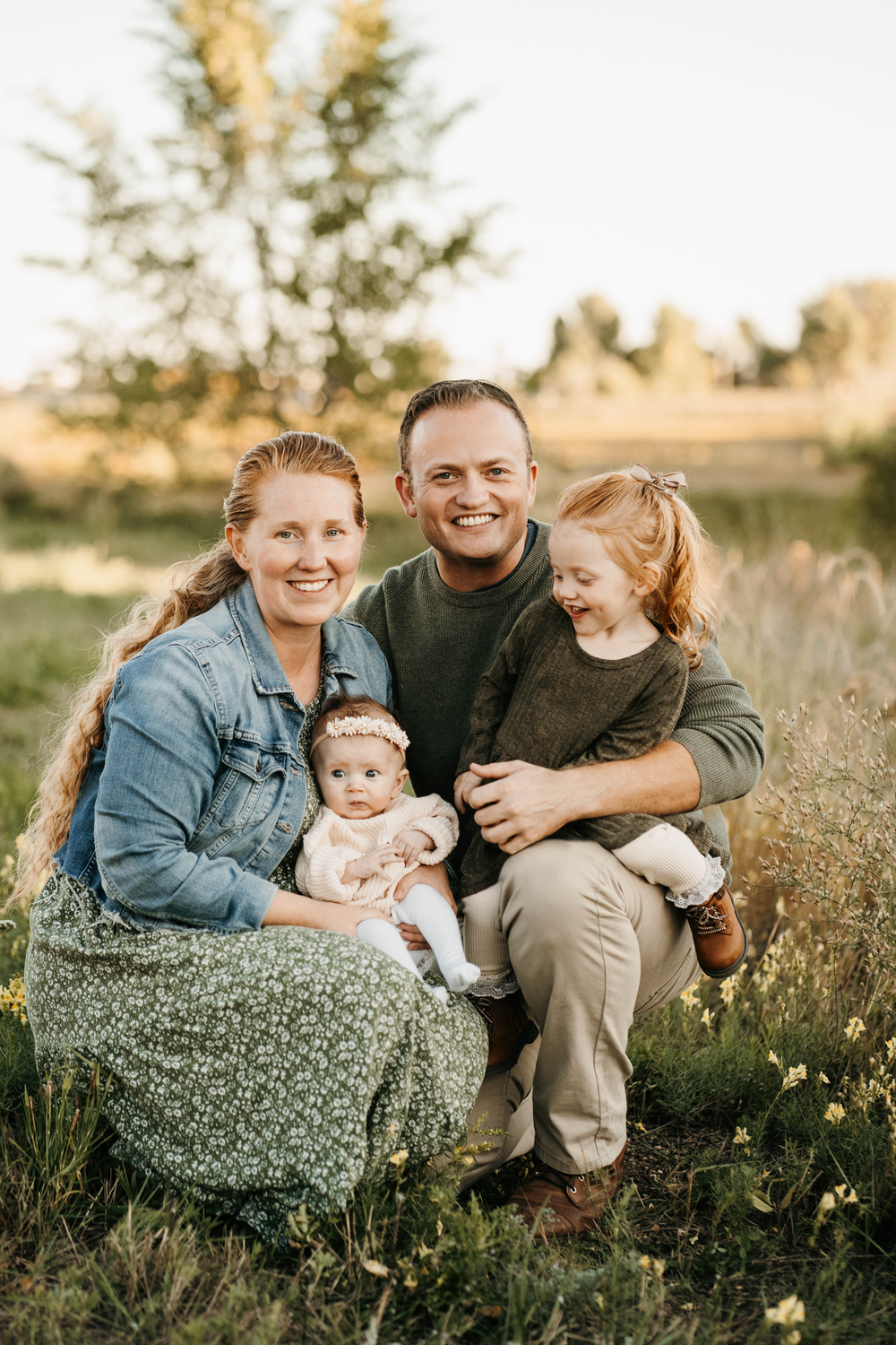 Happy family of four posing outdoors in a sunny meadow, parents smiling with two young daughters in a golden field.