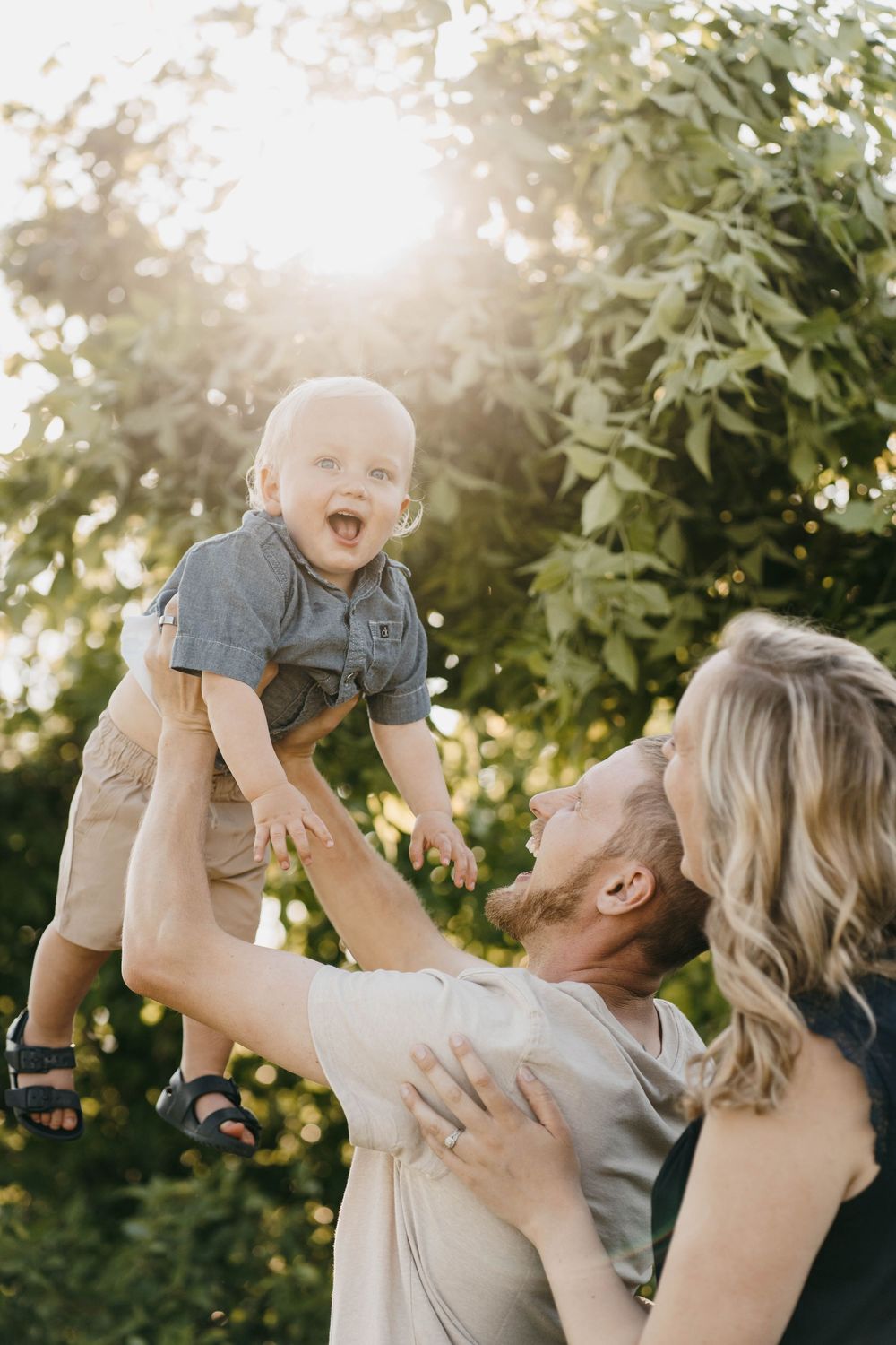 Happy baby laughing as parents lift him up outdoors in golden sunlight with lush green trees in background.