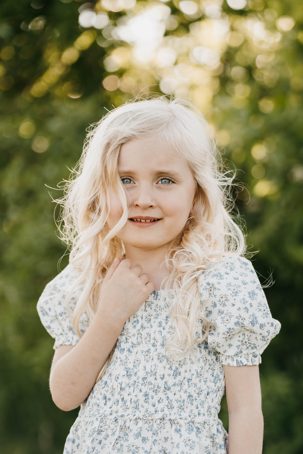 Young blonde girl in floral dress posing outdoors with green bokeh background in golden hour light.