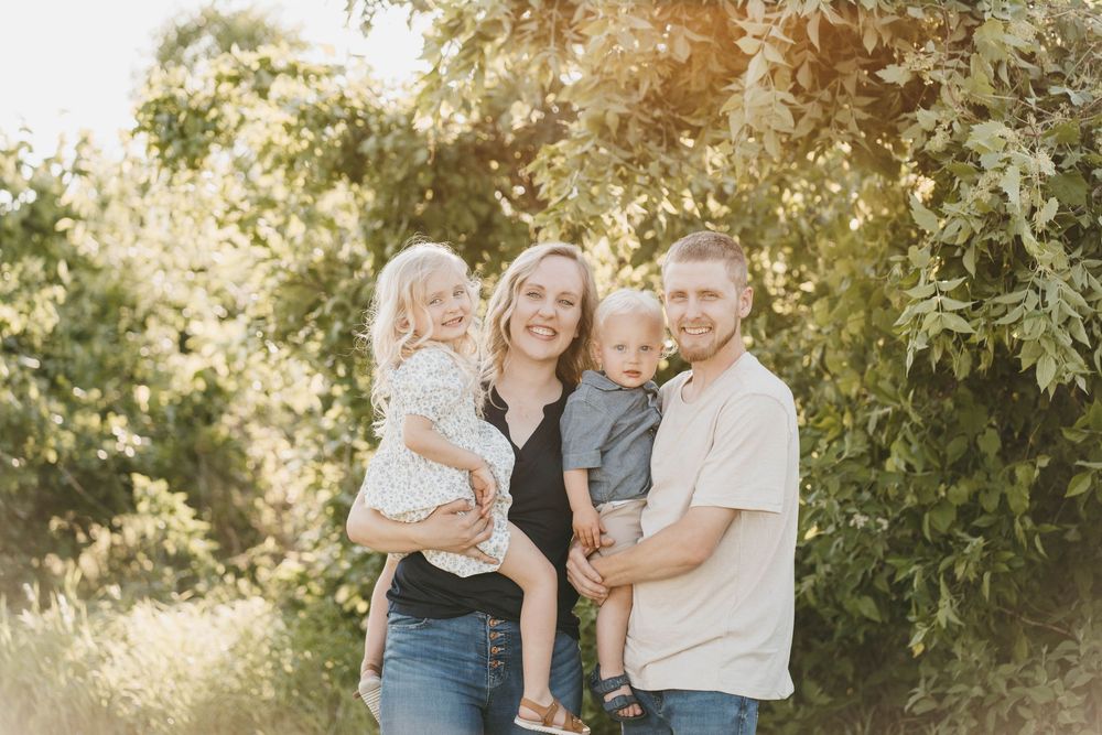 Happy family of four posing outdoors among lush green trees in warm sunlight, parents holding two young children.