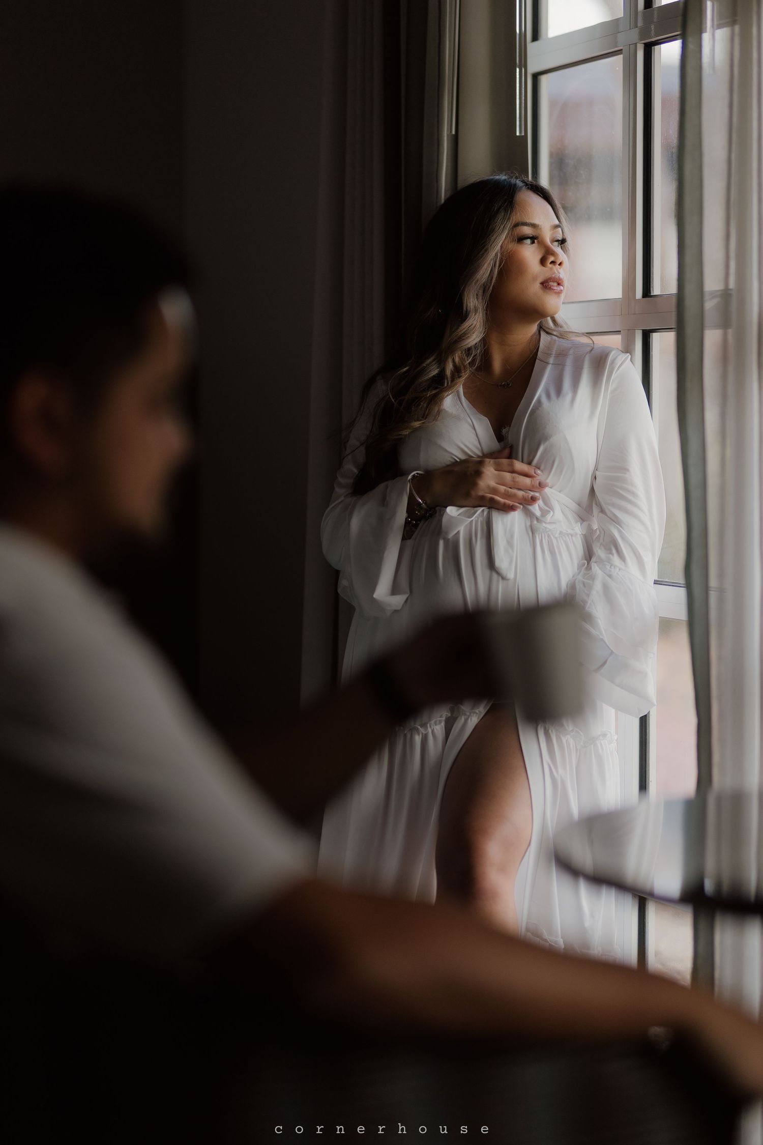 Artistic moody portrait of someone in a flowy white robe gazing thoughtfully through a window in soft natural light.