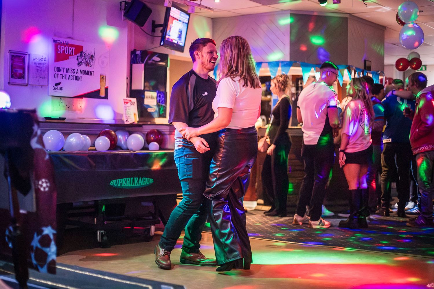 Man and a woman dance and smile at each other in a disco by a sports club bar.