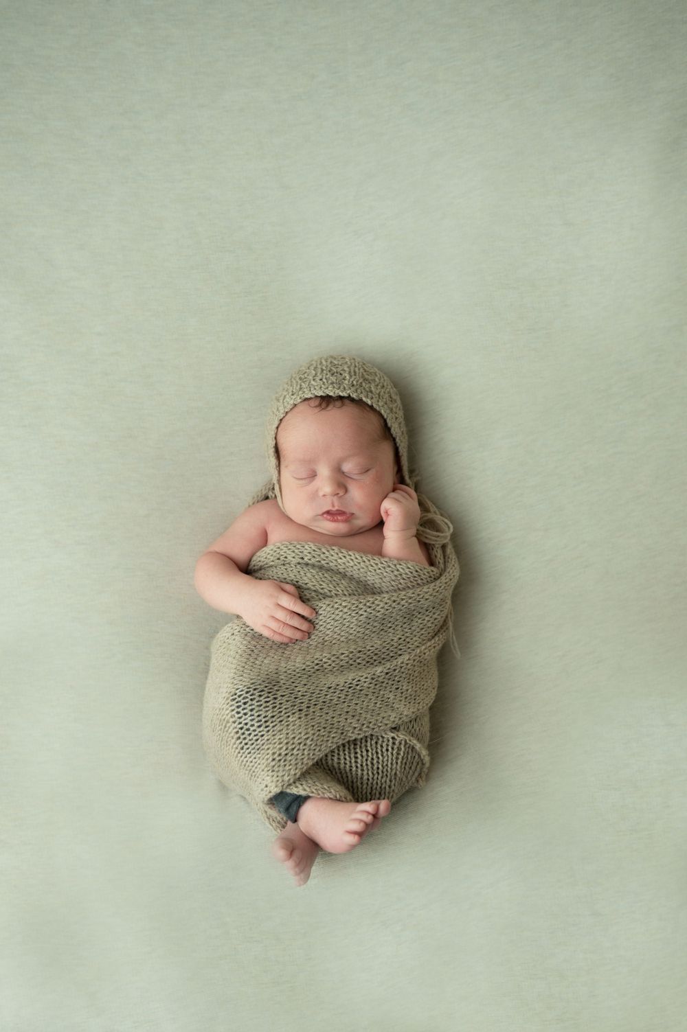 Sleeping newborn wrapped in knit blanket wearing bonnet on mint green backdrop.