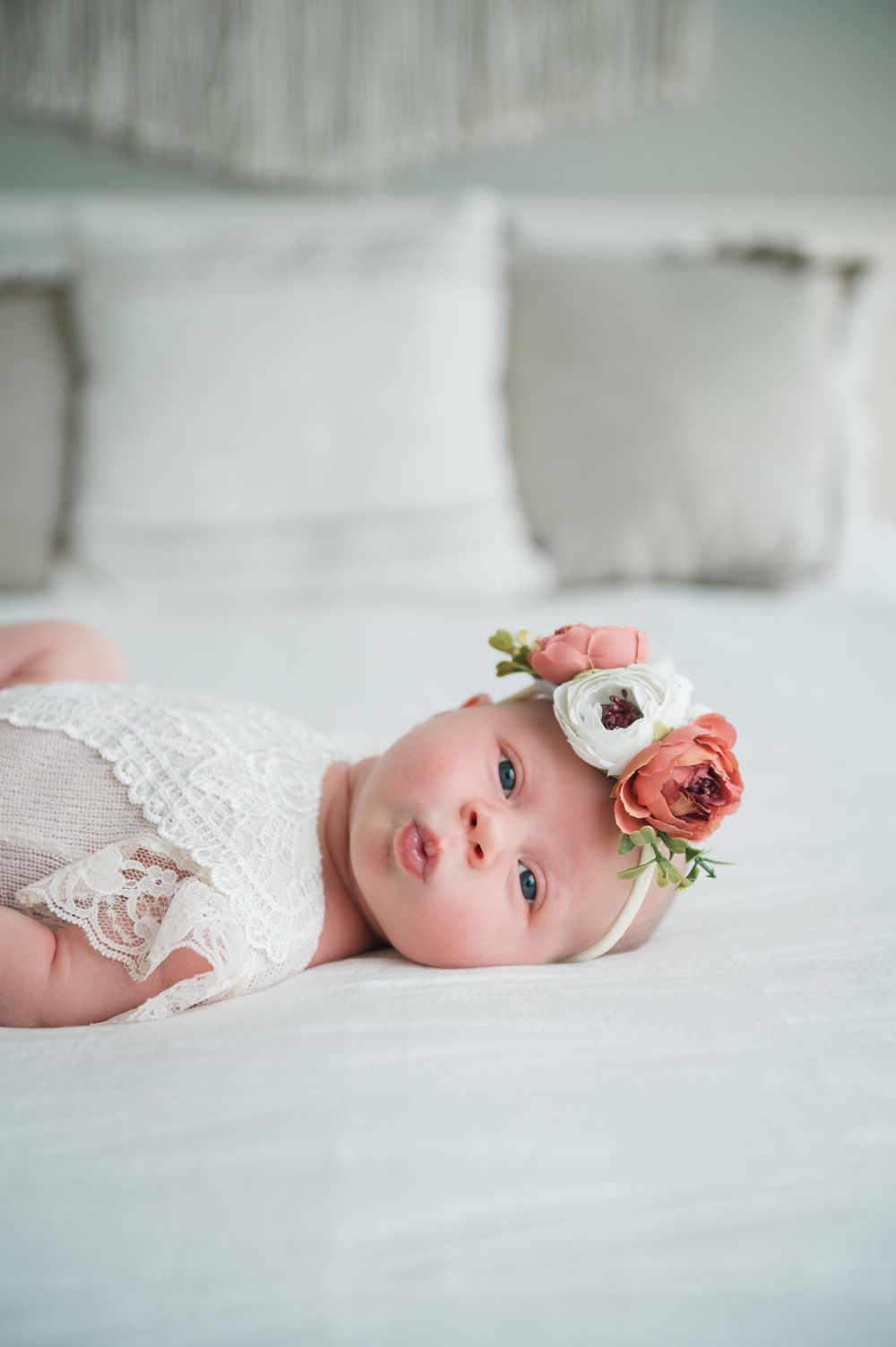 Baby in white lace outfit with floral crown rests on soft white bedding.