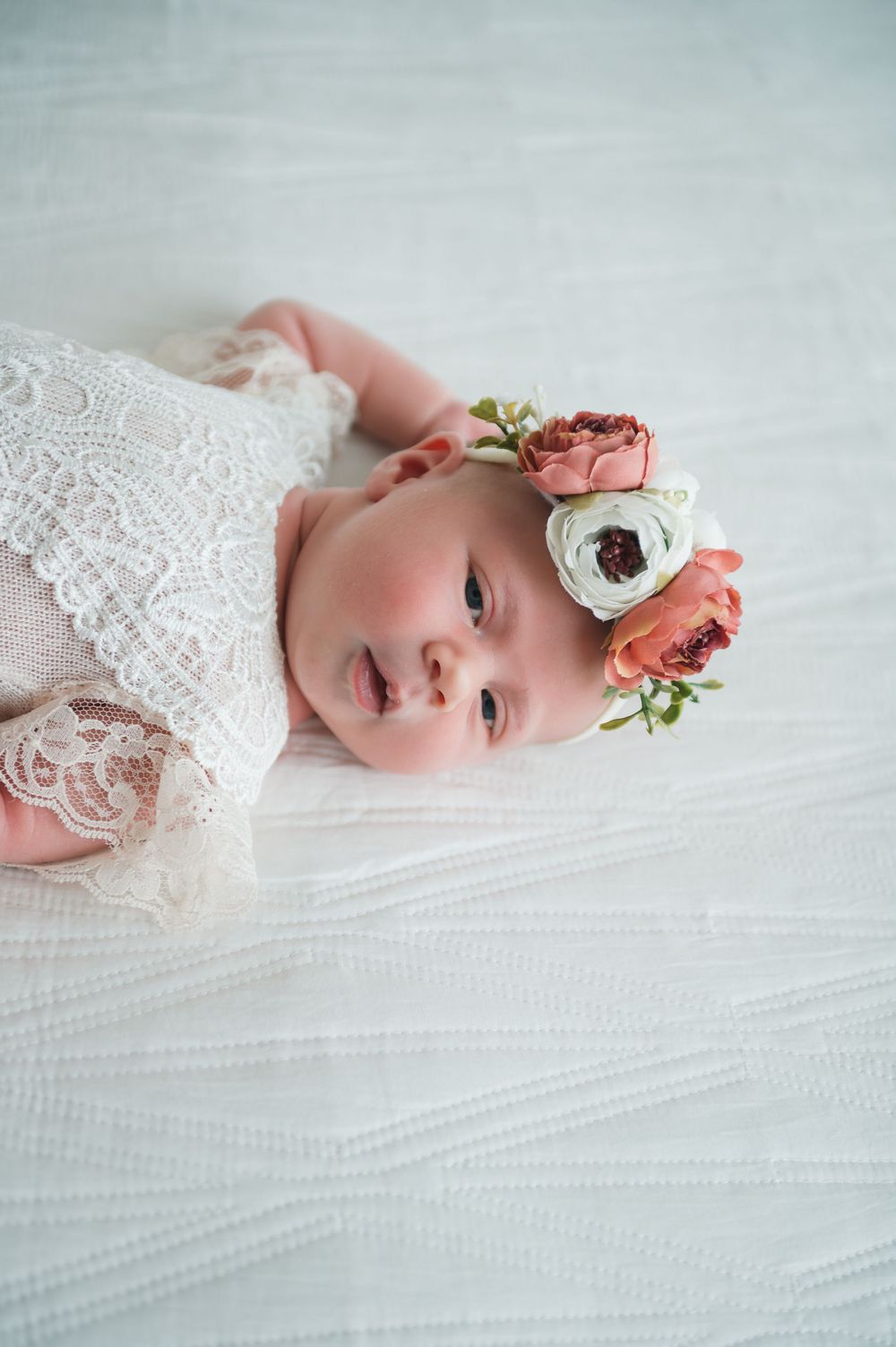 Infant wearing lace with pink and white floral crown on white textured background.