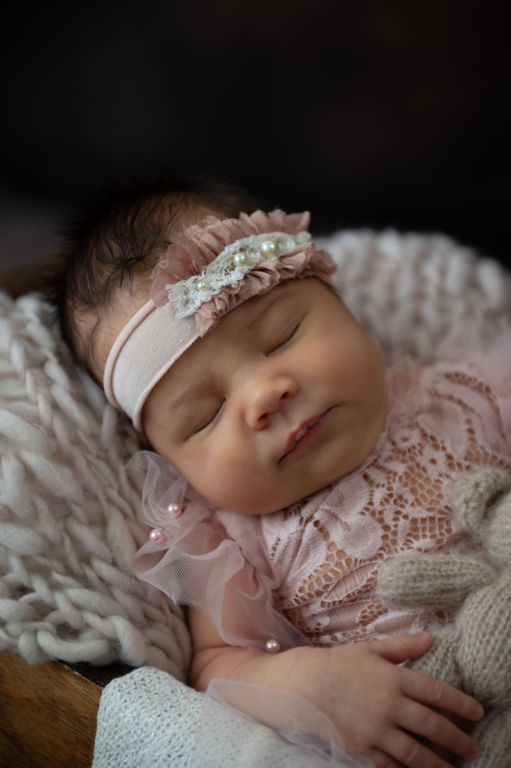 Newborn wearing pink lace dress and floral headband sleeps peacefully on textured white blanket.