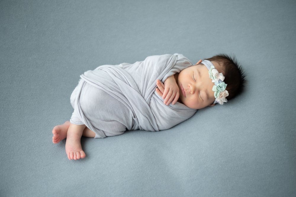 Sleeping infant in grey wrap photographed from above on light blue backdrop.