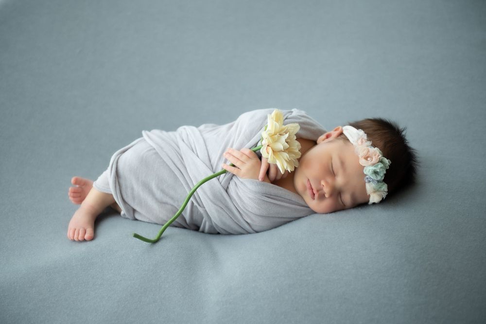 Newborn in grey swaddle with floral headband photographed on blue backdrop.