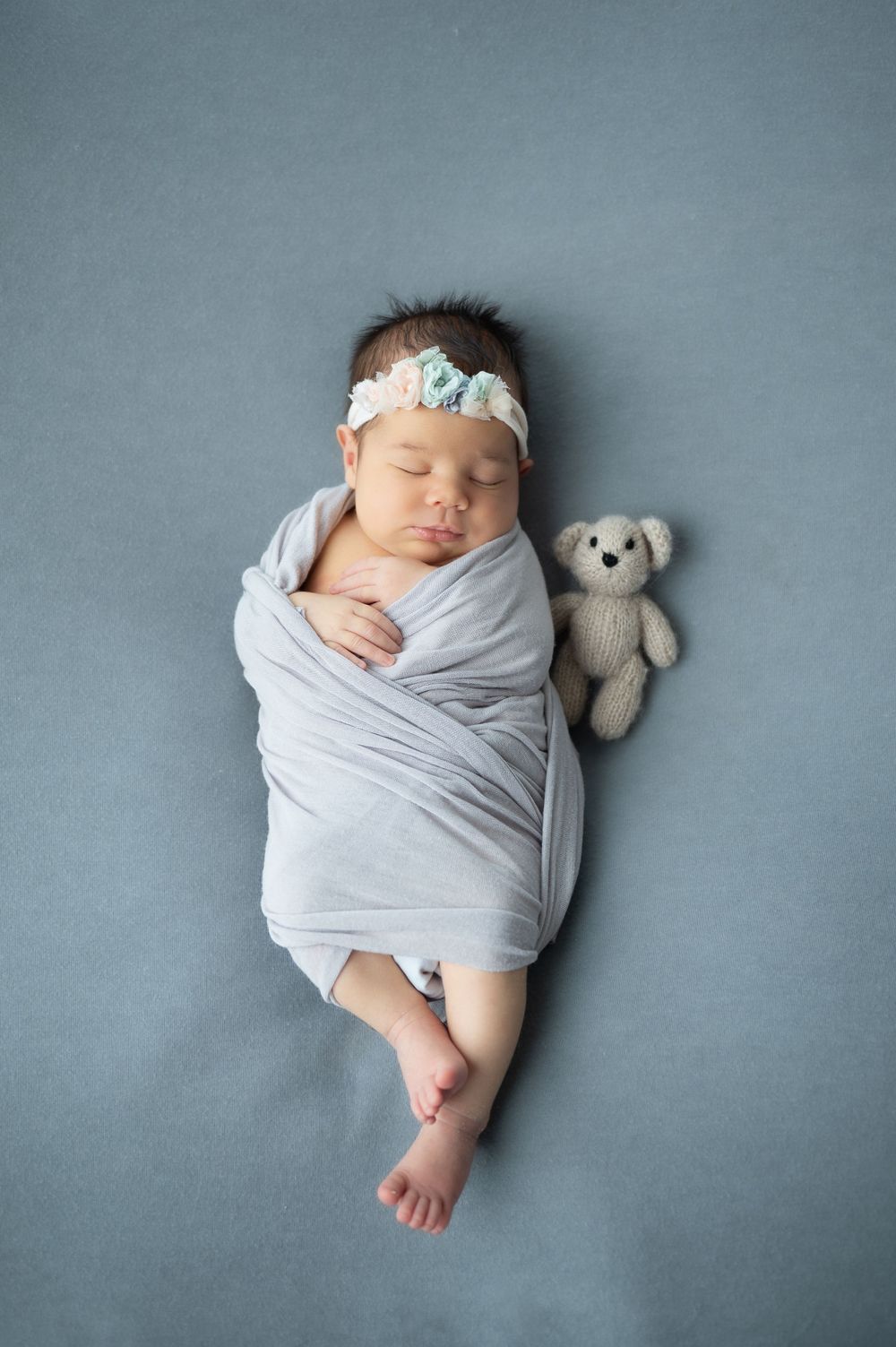 Sleeping baby wrapped in grey with small teddy bear companion on blue background.