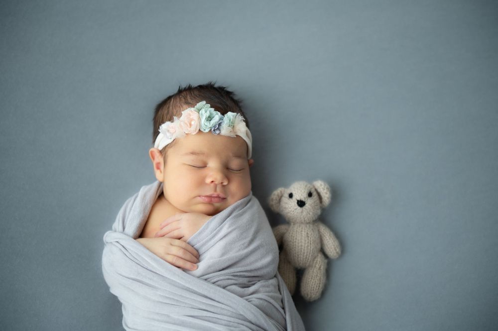 Peaceful newborn portrait with grey wrap and teddy bear against blue backdrop.