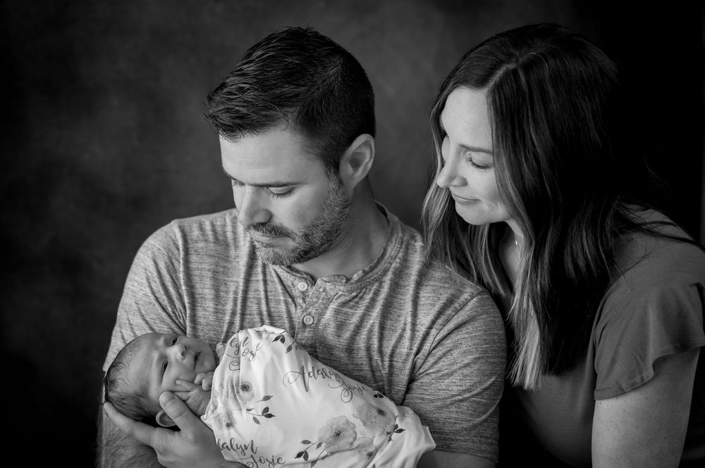 Black and white portrait of parents holding newborn baby against dark background.