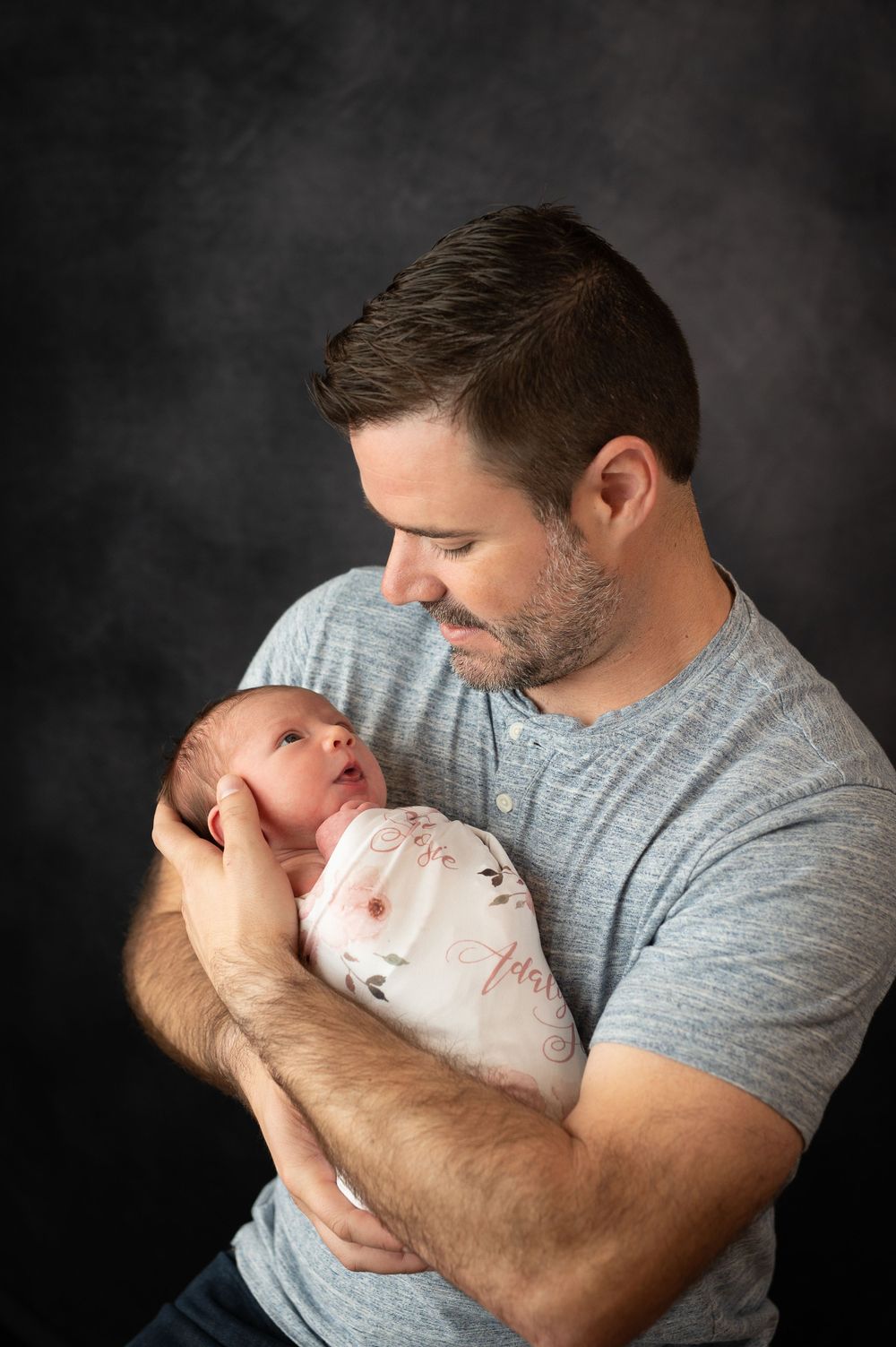 Father in grey t-shirt holding newborn baby against dark background.