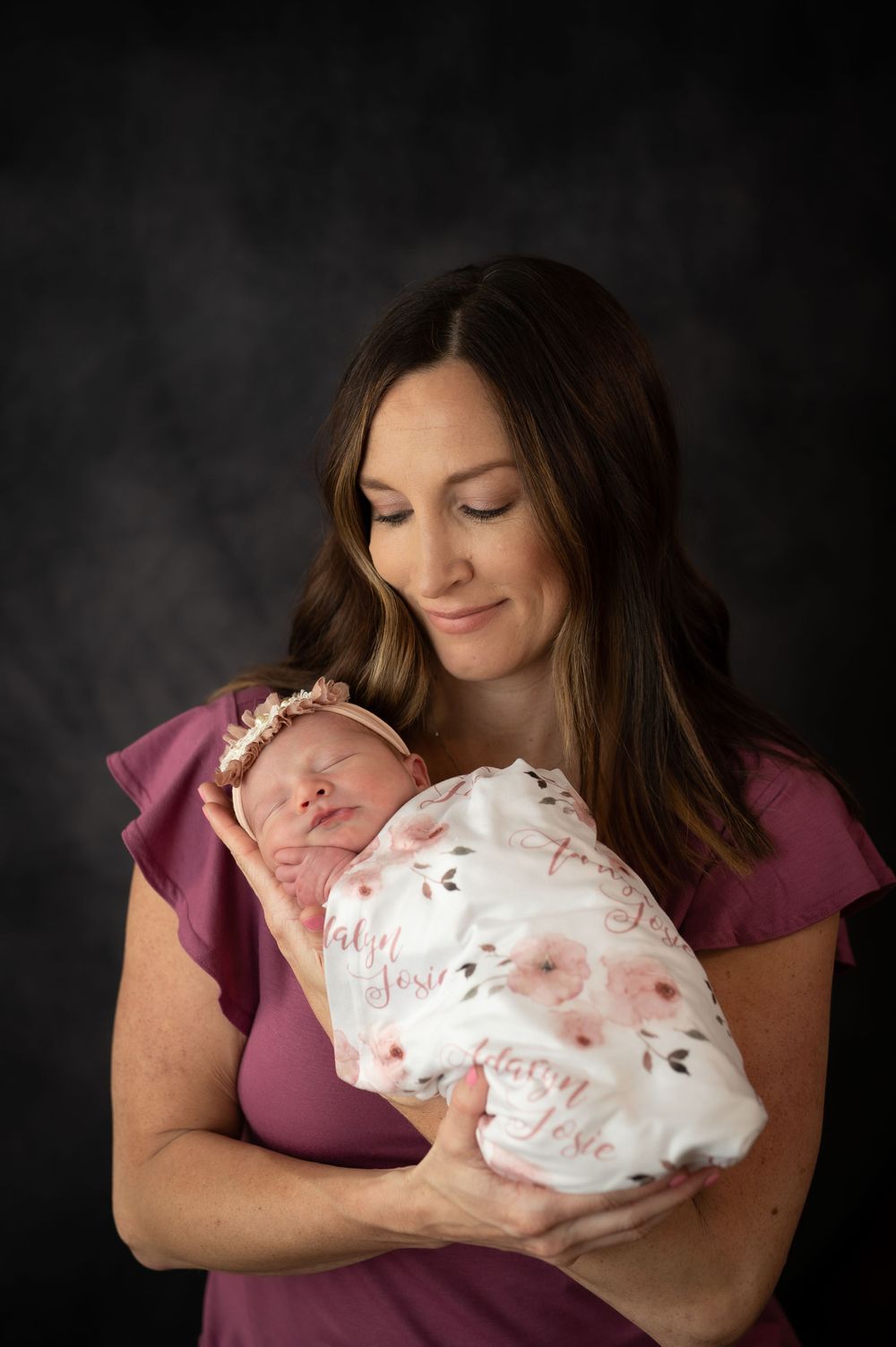 Mother in purple dress holding sleeping newborn wrapped in floral swaddle.