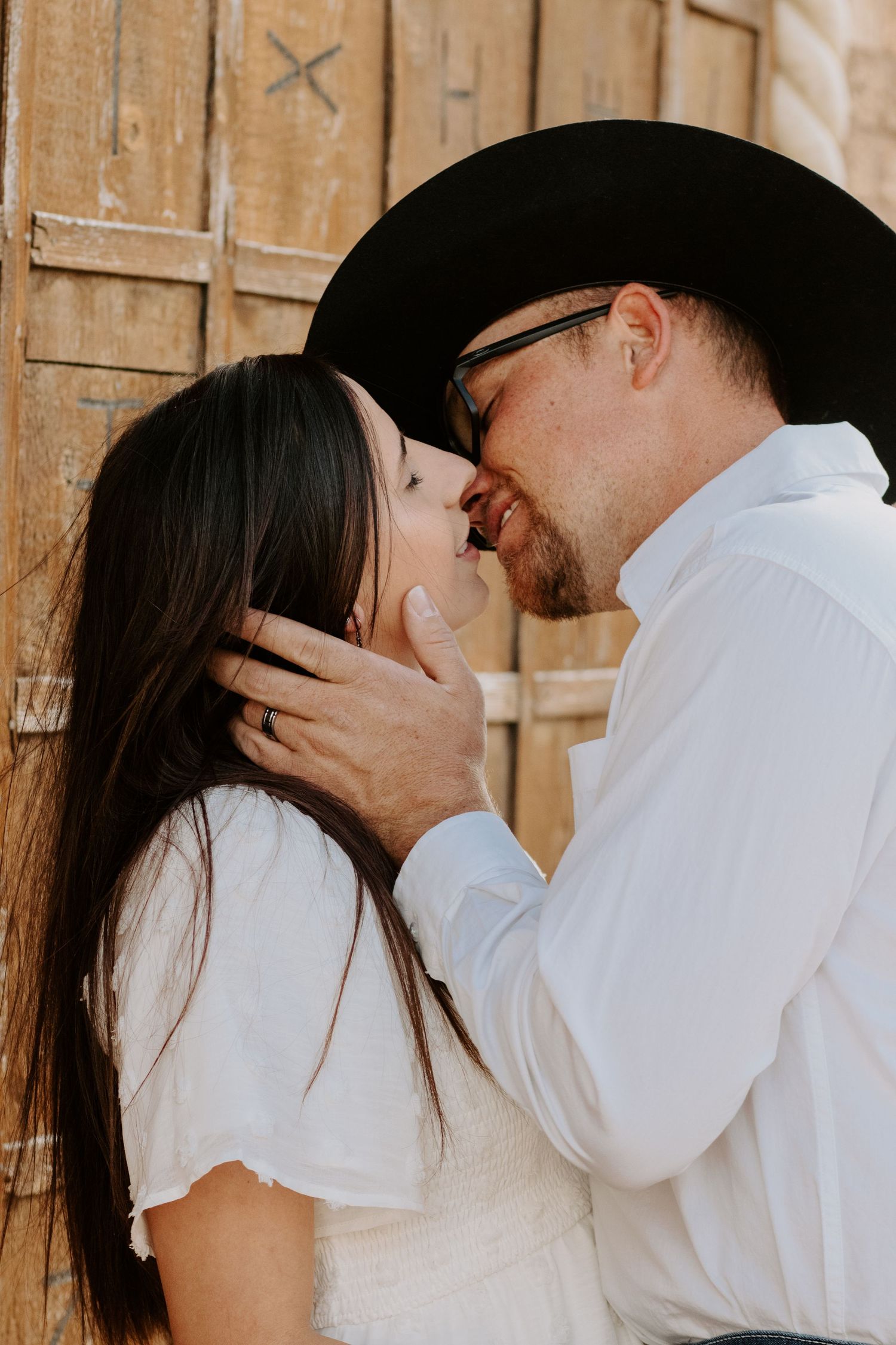 A couple in white outfits shares a romantic moment, man in black cowboy hat and glasses gently holding womans face.