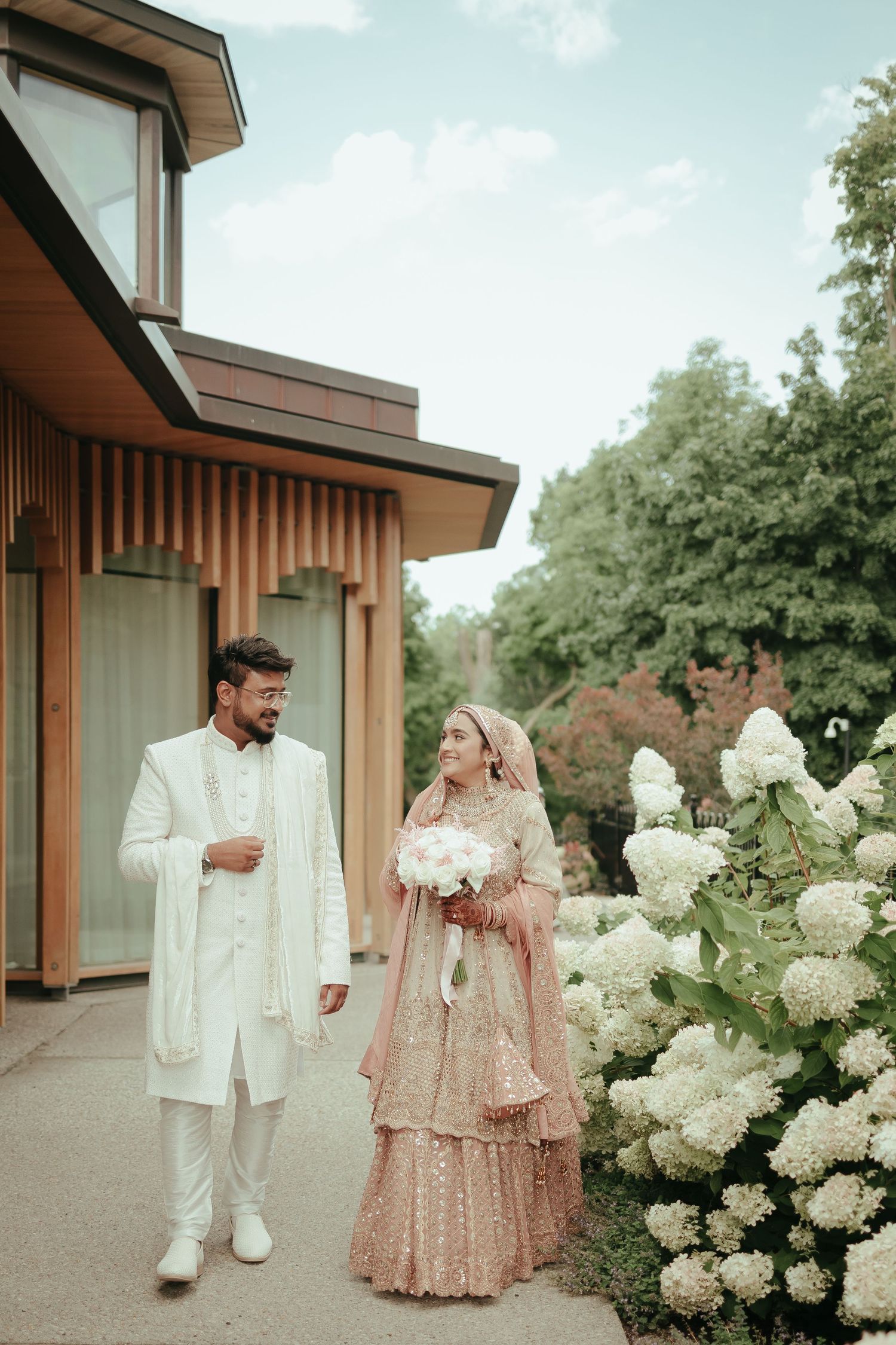 A wedding couple standing by blooming white hydrangea bushes outside a modern building.