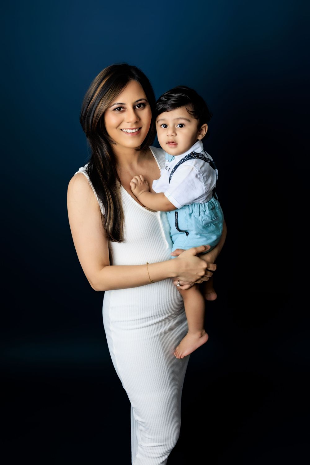 A parent and child pose against a dark blue background, with the parent wearing a white dress and holding the toddler.