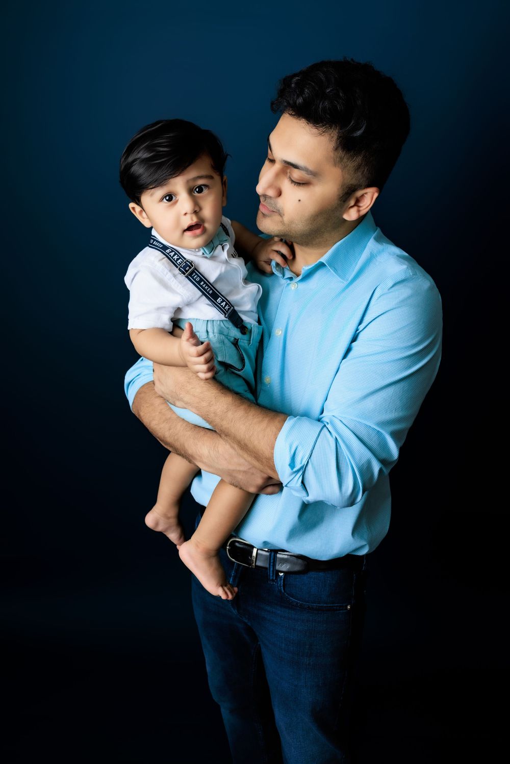 A parent in a light blue shirt holds a small child wearing white against a dark background in a studio portrait.