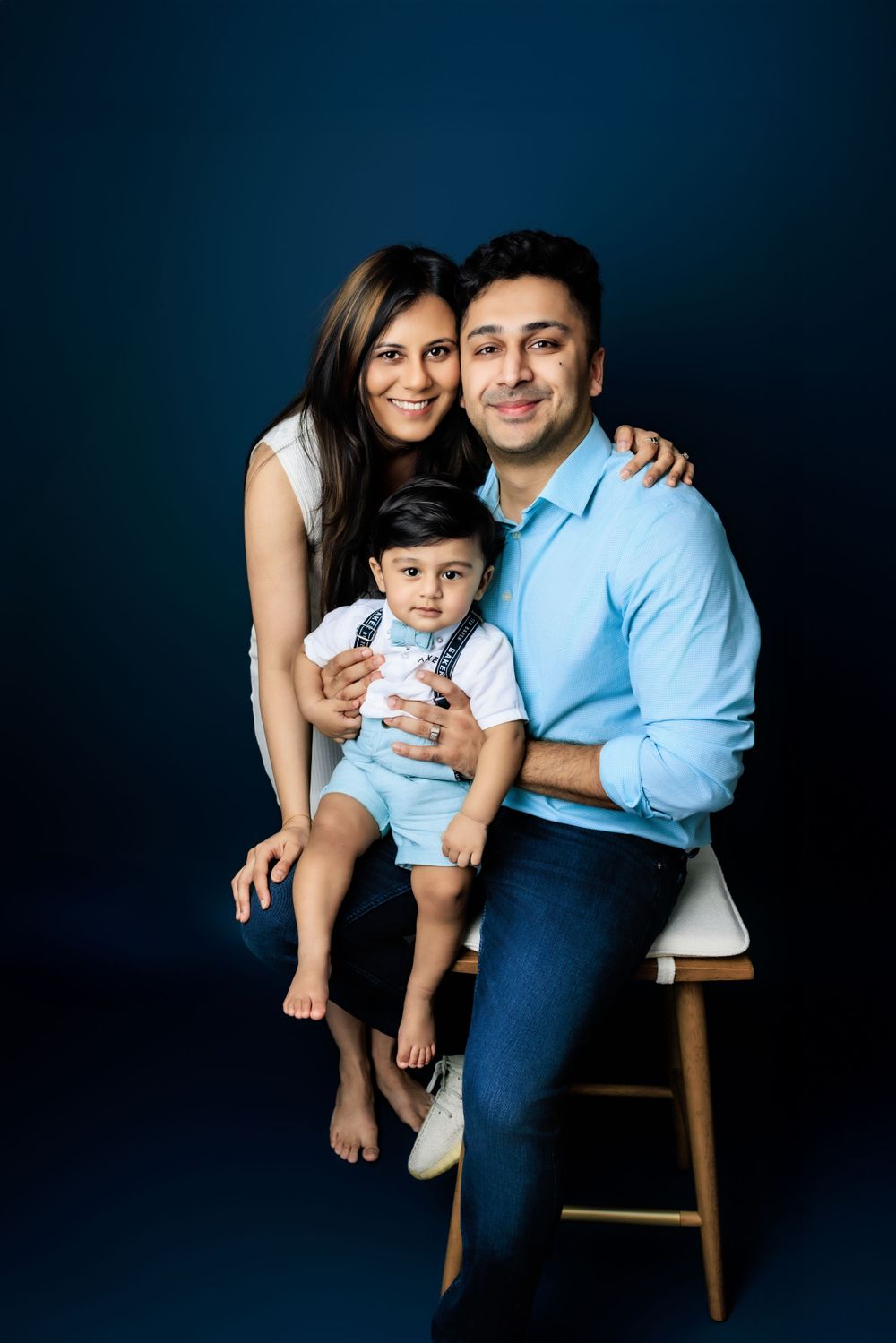 A family of three poses together on a wooden stool against a dark blue backdrop for a studio portrait.