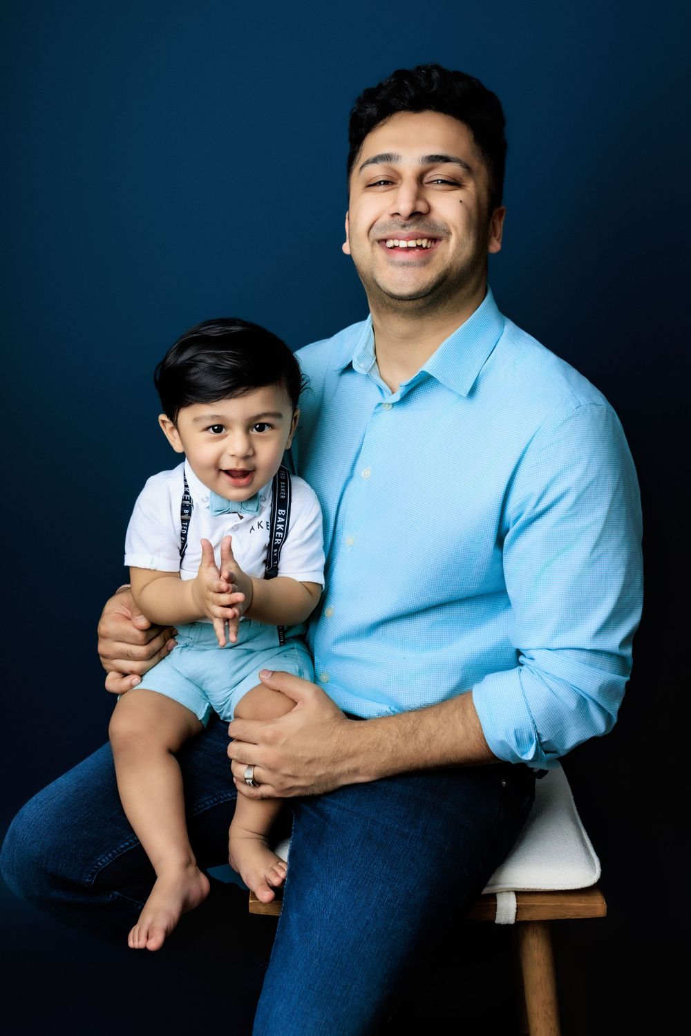 A parent and child share joyful smiles while sitting together on a wooden stool against a blue background.