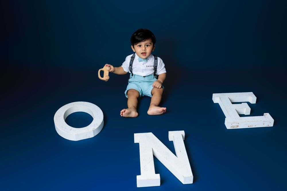 Baby in blue overalls sits among large white letters spelling ONE against a dark blue background.