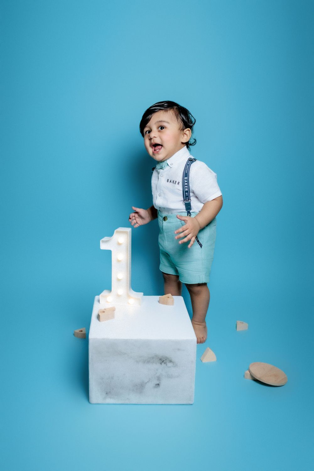 Small child in white shirt and blue shorts celebrating first birthday on white cube pedestal against blue background.