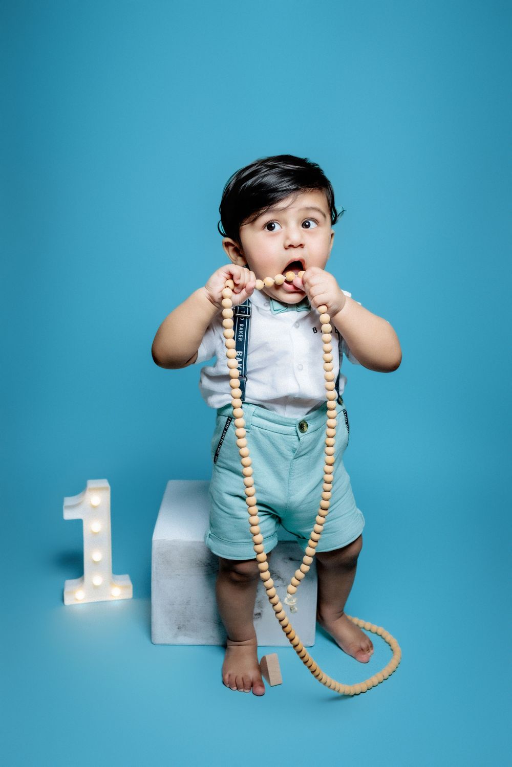 A small child in overalls stands on a white block next to a light-up number 1, playing with a long beaded necklace.