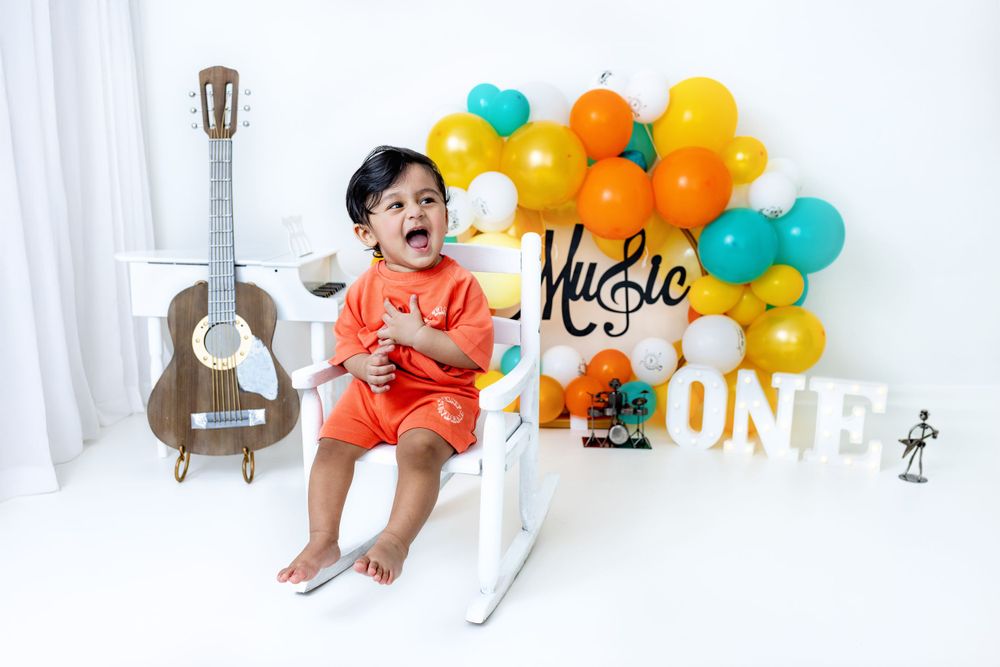 A baby sits near a guitar and colorful orange and teal balloons with birthday party decorations.