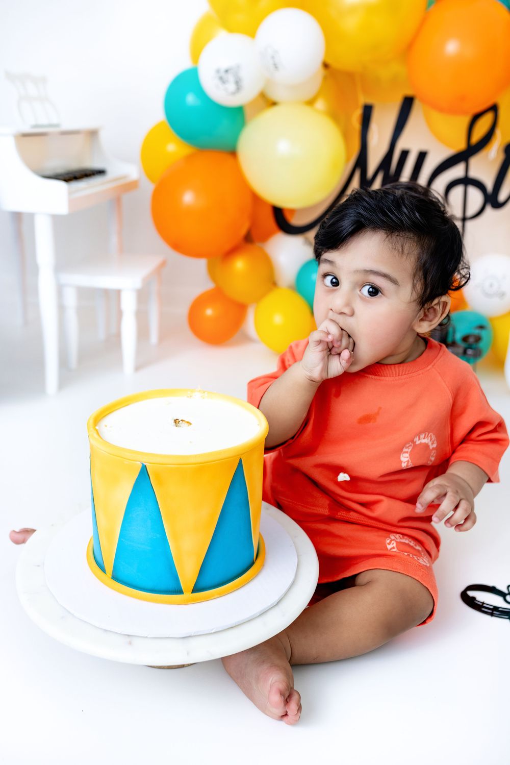 A small toddler in orange clothing sits next to a drum-shaped cake with colorful balloon decorations.
