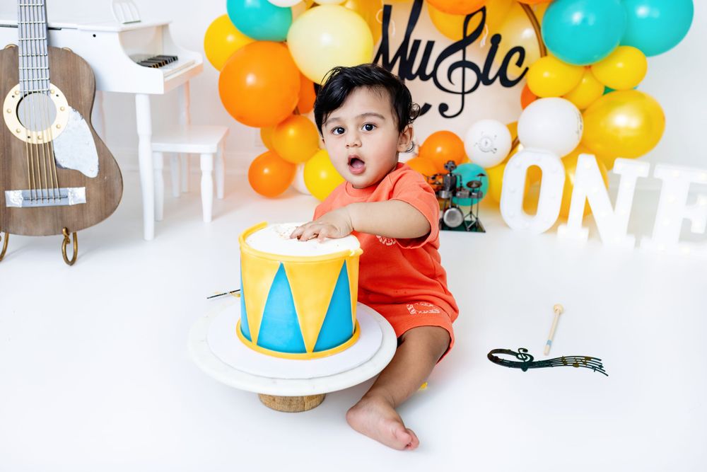 Baby in orange outfit hits drum during music-themed first birthday party with colorful balloon decorations.