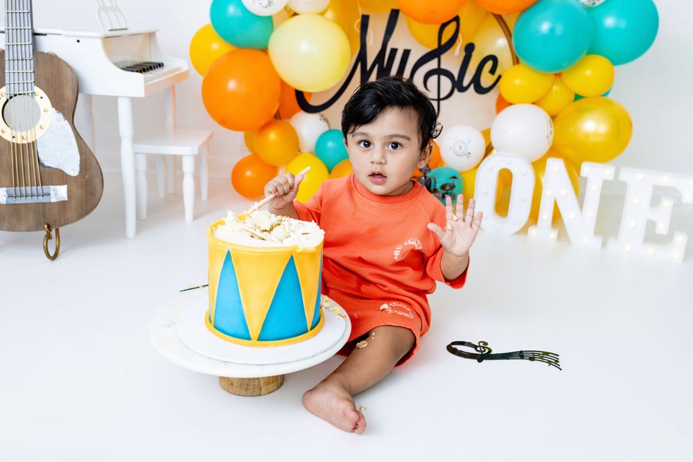 A baby sits by a first birthday cake with colorful balloons, drum decorations and a guitar in the background.
