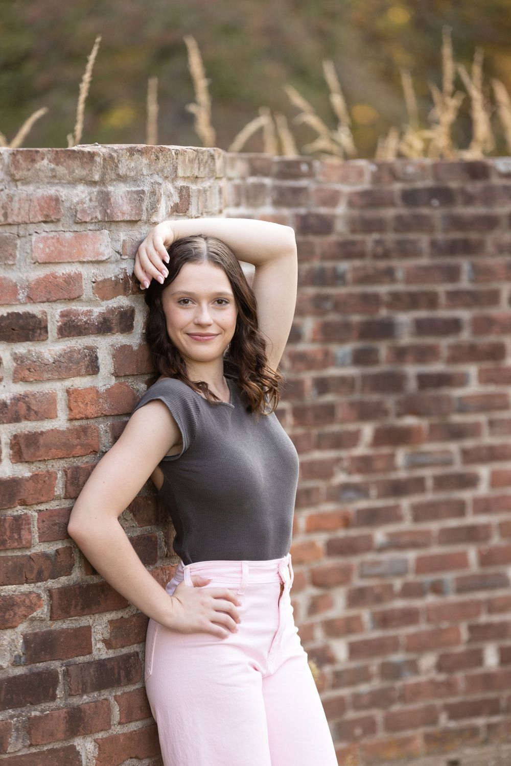 A fashionable outfit featuring a grey top and pink skirt is modeled against a rustic brick wall.