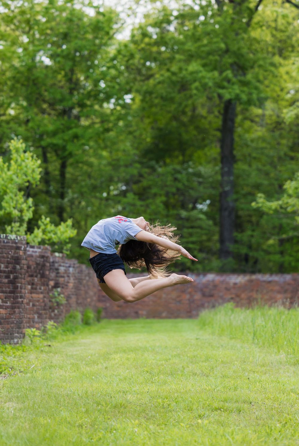 A dancer performs a graceful horizontal leap across a grassy lawn in front of a brick wall on a sunny day.