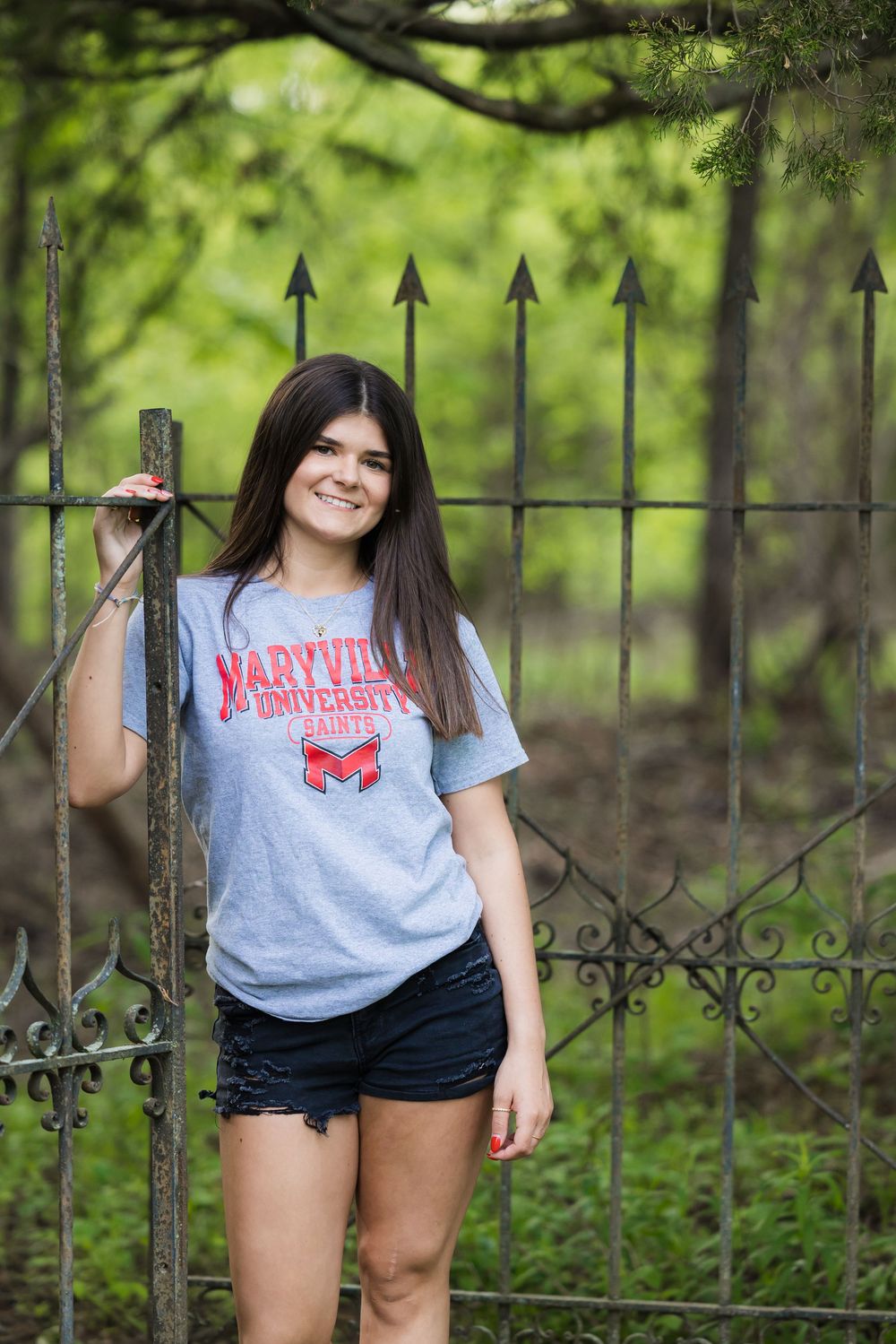 A student wearing a university t-shirt poses casually by a decorative iron fence in a green outdoor setting.