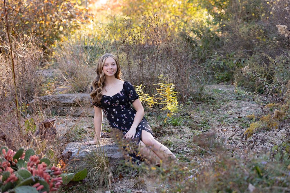 A serene autumn countryside photoshoot captures golden sunlight filtering through trees near a stone wall at sunset.
