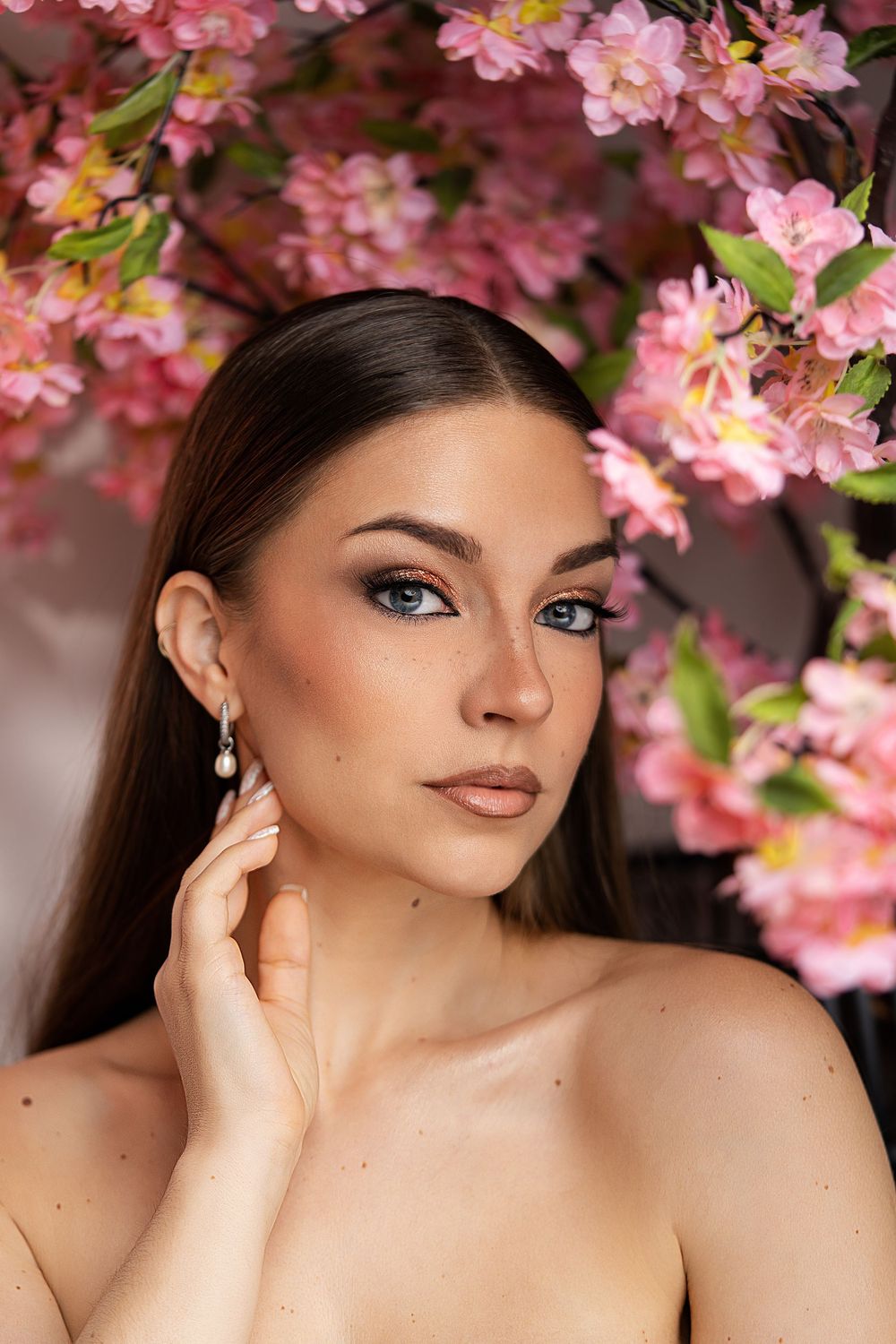 Young woman with blue eyes and pearl earring poses gracefully among pink cherry blossoms in a beauty portrait.