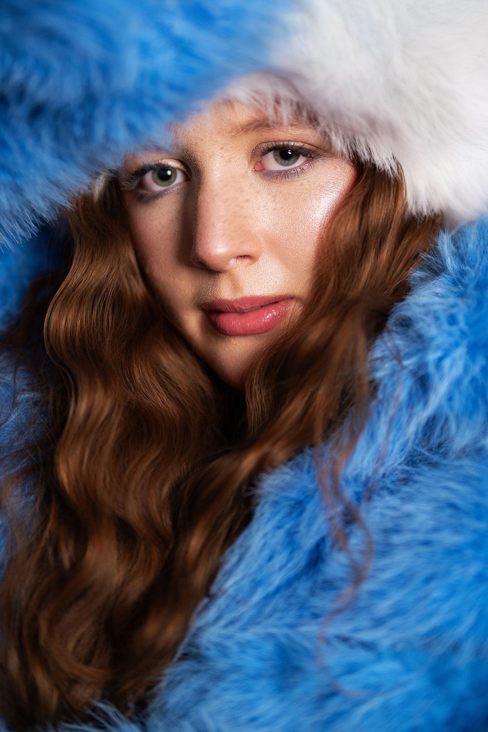 Young woman with freckles and wavy red hair wearing a fluffy blue and white fur hood coat.