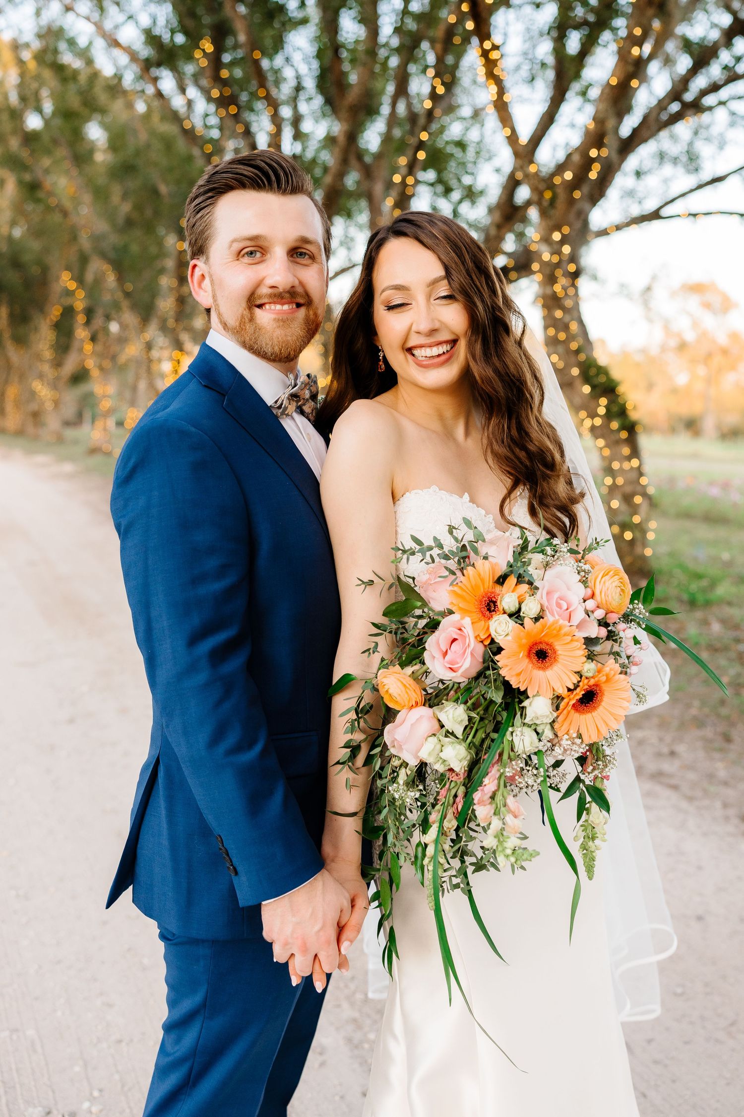 Bride holding boho bouquet while groom embraces her from behind against neutral white background.
