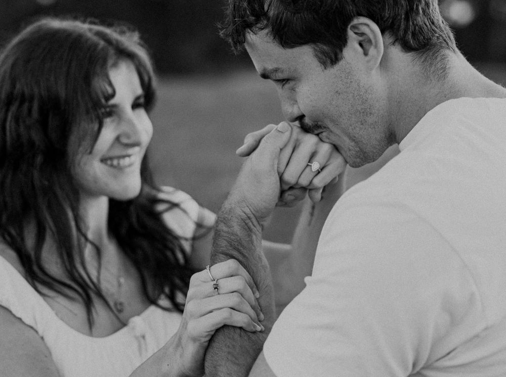 Black and white photo of a romantic couple, man kissing womans hand while she smiles tenderly outdoors.