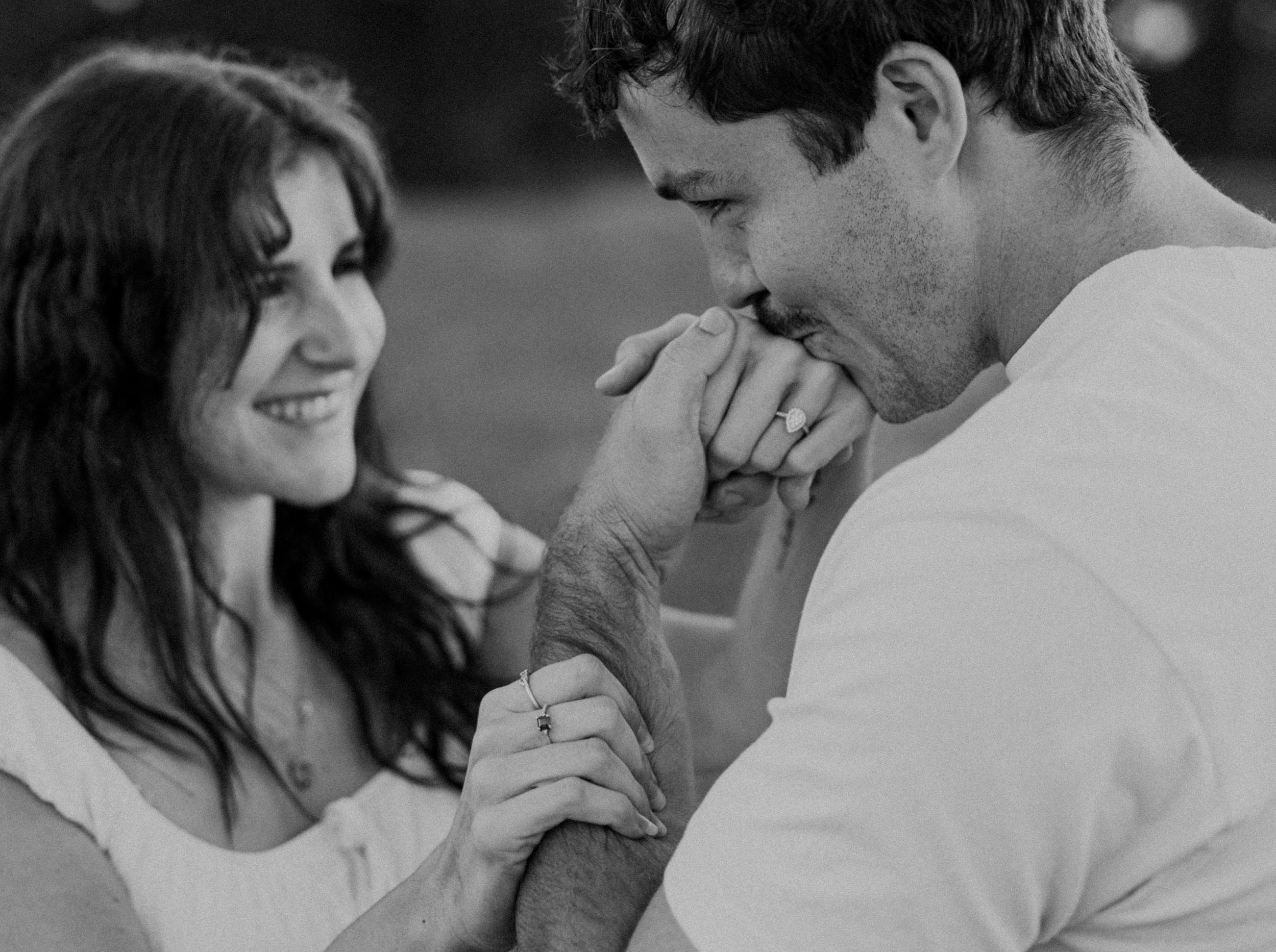 Black and white photo of a romantic couple, man kissing womans hand while she smiles tenderly outdoors.