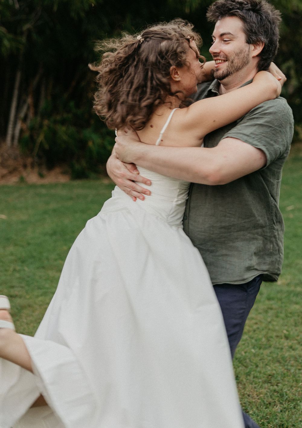 Joyful couple embracing outdoors, bride in white wedding dress being lifted by groom in green shirt on grass.