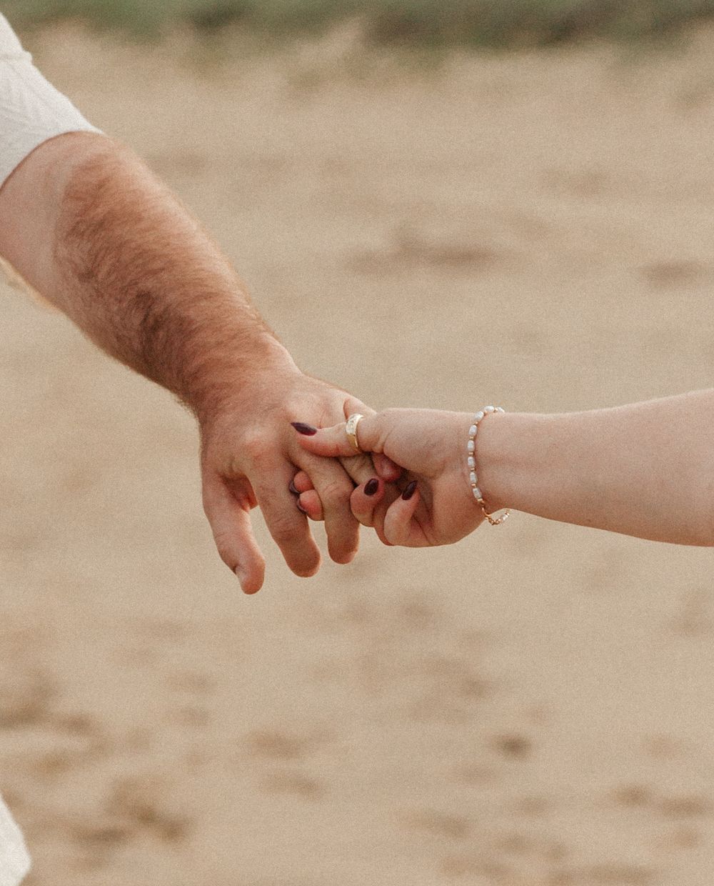 Close-up of couple holding hands on sandy beach, woman wearing gold rings and pearl bracelet.