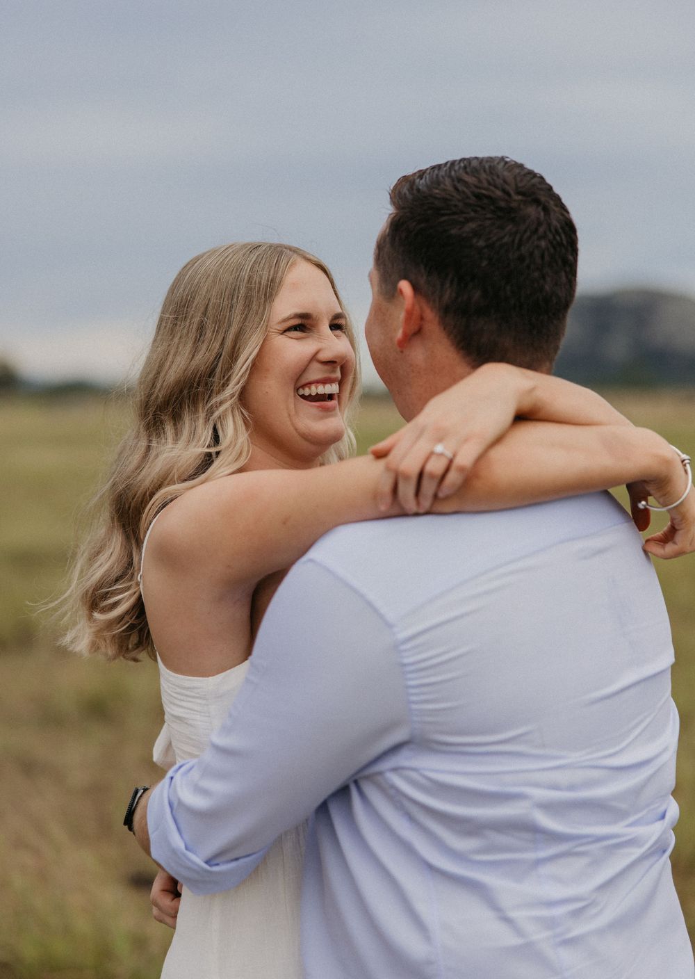 A joyful couple embraces in an open field, the smiling blonde woman showing off her engagement ring.