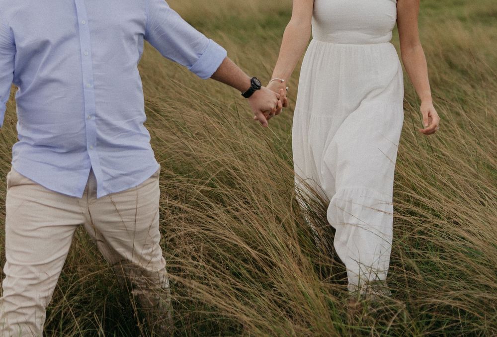 Couple holding hands walking through tall grass field, man in blue shirt and woman in white dress.