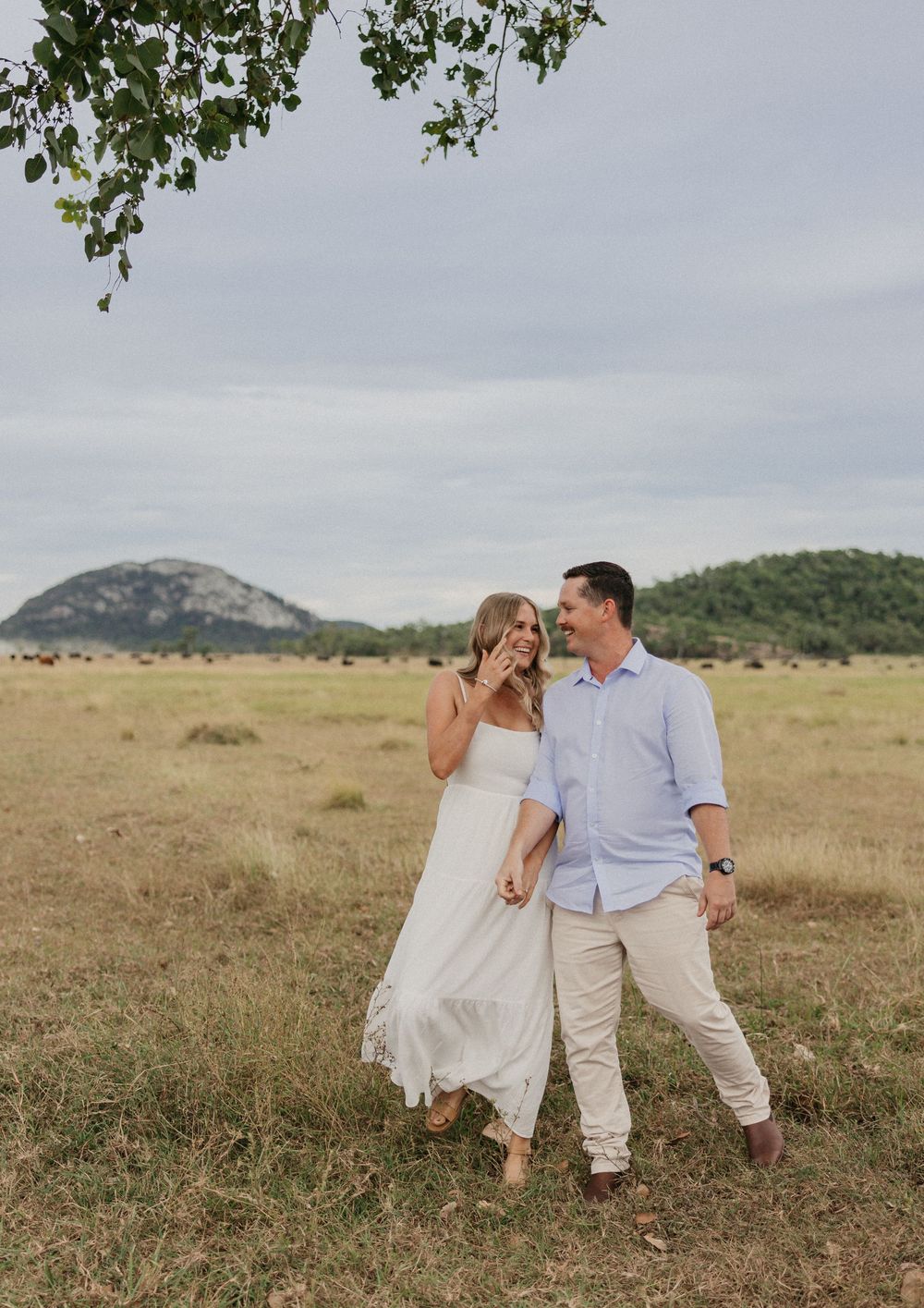 Couple laughing and holding hands in a grassy field with a scenic hill backdrop during an outdoor engagement photo session.
